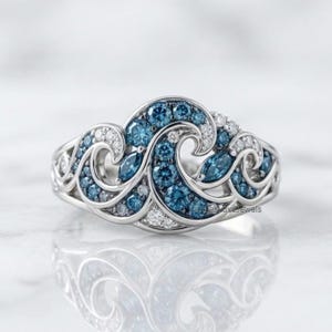 Può includere: Un anello color argento con un design ispirato alle onde, incastonato con molteplici pietre blu rotonde e taglio marquise e piccole pietre chiare. La fascia metallica si curva in forme fluide e vorticose che ricordano le onde del mare.