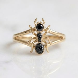 May include: A gold ring featuring an ant-shaped centerpiece. The insect design is crafted from polished gold and accented with multiple small, round black gemstones set into the head, thorax, and abdomen sections of the ant's body structure.