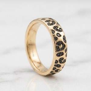 May include: A gold ring with a leopard print design. The ring has a band with black gemstones arranged in a pattern similar to leopard spots. The ring is displayed on a white marble surface.