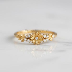 May include: A gold ring with a floral design, featuring a central daisy and surrounding leaves. Small, clear gemstones are set around the flower and leaves. The ring is on a white surface, with the text "VenuesLuxejewels" visible.