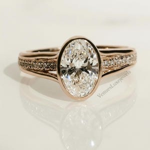 May include: Rose gold engagement ring with an oval-cut diamond center stone. The band features a row of smaller diamonds. The ring is set against a white background, with the text "VenuesLuxeJewels" visible.