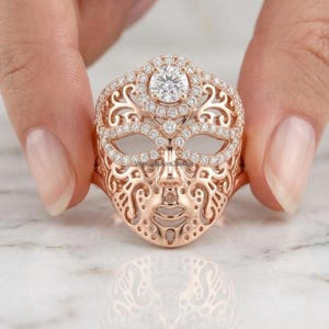 Könnte beinhalten: Ein Ring aus Roségold mit einem maskenförmigen Mittelstück, das mit kleinen, pavé-gefassten weißen Steinen besetzt ist. Das Maskendesign weist komplizierte filigrane Ausschnitte und einen runden, brillantgeschliffenen weißen Stein auf.
