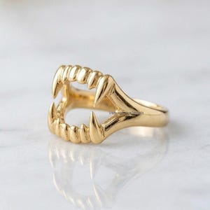 May include: A gold-toned ring shaped like a set of open vampire fangs, featuring a smooth, polished metallic finish. The design includes pointed canine teeth and smaller incisors, creating a continuous band that encircles the finger.