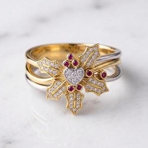 Op de afbeelding: Een ring met meerdere banden en een centraal hulstbladmotief van geelgoud met pavé-gezette witte diamanten. Het ontwerp bevat een hartvormig diamantcluster in het midden en kleine ronde rode robijnen. De tekst VenuesLuxeJewels is zichtbaar.