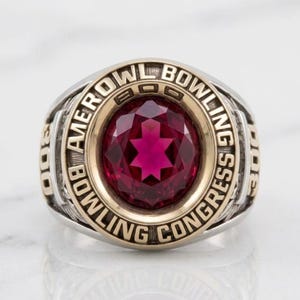 May include: A two-tone ring with a large, faceted, ruby-red gemstone at its center. The ring has gold-colored accents and the words "AMEROW BOWLING CONGRESS" are inscribed around the gemstone. The ring is a classic style.