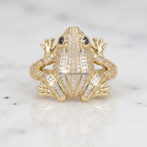 Puede incluir: Un anillo de oro con forma de rana, decorado con pequeñas piedras blancas redondas y piedras blancas en corte baguette. La rana tiene dos piedras oscuras y redondas como ojos. La banda del anillo tiene textura que imita las patas de una rana.