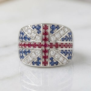 May include: A wide silver band ring featuring a Union Jack flag design created with inlaid round-cut red rubies, blue sapphires, and clear white diamonds. The gemstones are arranged in a cross and diagonal pattern across the face of the ring.
