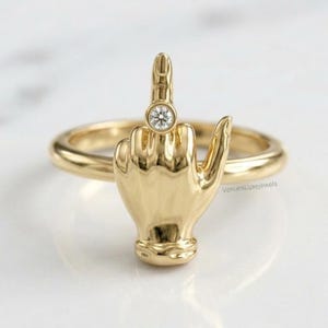 Op de afbeelding: Een gouden ring met een gebeeldhouwde hand die een middelvingergebaar maakt, met een kleine ronde heldere edelsteen gezet op het gewricht van de middelvinger. De tekst VenuesLuxeJewels is zichtbaar op de zijkant van de ringband bij de hand.