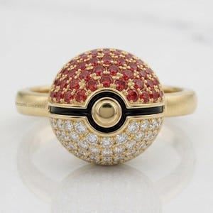 May include: A gold ring featuring a spherical centerpiece designed like a Poké Ball. The top half is encrusted with red gemstones, the bottom half with clear white stones, and a central black band with a gold circular accent in the middle.