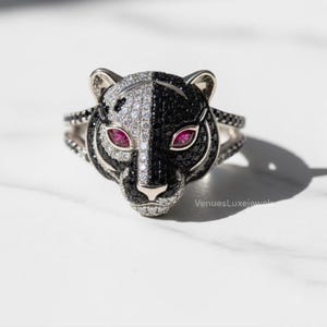 May include: A silver ring with a panther head design. The panther's face is split, with one side covered in white stones and the other in black stones. The eyes are small, red gems. The ring has a split band with black stones.