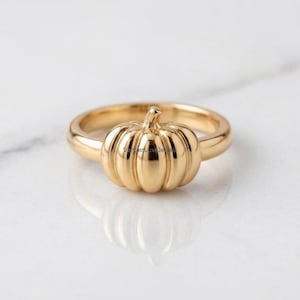 May include: A gold ring featuring a central pumpkin-shaped charm with vertical ridges. The band is smooth and circular, polished to a high shine. The metal surface reflects light, highlighting the rounded contours of the pumpkin design.