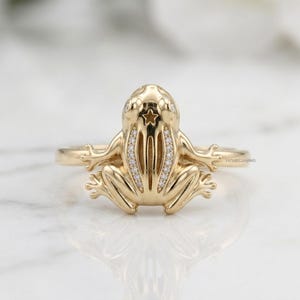 May include: A gold ring featuring a sculpted frog design on the front. The frog has a small star cutout on its head and two rows of tiny clear stones embedded along its back. The band is smooth and polished, showcasing a detailed amphibian form.