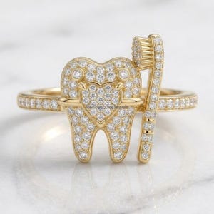 Puede incluir: Un anillo de oro con un motivo central en forma de diente y un diseño de cepillo de dientes, ambos incrustados con numerosas piedras blancas pequeñas. La banda también está pavimentada con piedras blancas y el texto venuestluxejewels es visible.
