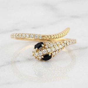 May include: A gold-colored snake ring with a coiled design. The ring features a head with two black oval stones and numerous small white stones. The band is adorned with small white stones and a textured pattern.
