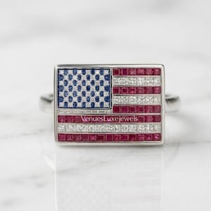 May include: A silver ring featuring a rectangular design of the American flag. The flag design includes blue sapphire stones for the stars, red rubies, and white diamonds for the stripes. The text "VenuesLuxeJewels" is visible.