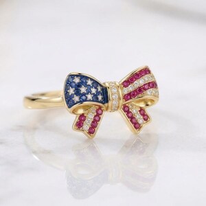 May include: A gold ring featuring a bow-shaped centerpiece adorned with blue stones, red stones, and small clear stones. The blue section includes star-shaped accents, while the red section features striped patterns, all set in a polished gold band.