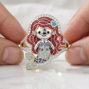 May include: A gold ring featuring a mermaid character with long red hair set with red stones, a blue stone heart hair accessory, and a tail decorated with light and dark blue stones. The border is lined with small white stones.
