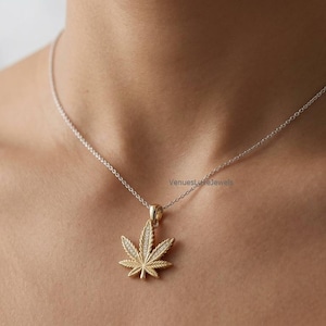 May include: A gold-toned pendant necklace featuring a cannabis leaf design encrusted with small clear stones. The pendant hangs from a delicate, thin silver-toned chain. The text VenuesLuxeJewels is visible near the top of the pendant.