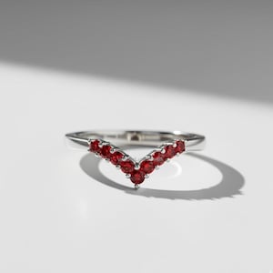 Peut inclure: Une bague en argent avec un design en forme de V. Elle est ornée de petites pierres précieuses rouges rondes. Les pierres sont serties de près, créant une ligne continue de couleur. La bague est présentée sur une surface blanche.