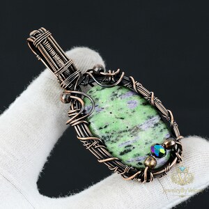 Puede incluir: Un colgante ovalado con una piedra moteada en verde y morado, engastada en un intrincado alambre de cobre oscuro. El diseño incluye una anilla enrollada en la parte superior y pequeñas cuentas metálicas integradas en el marco de alambre.