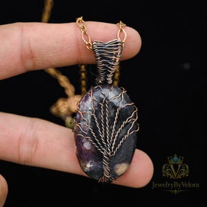 Puede incluir: Un collar con colgante hecho a mano con una piedra ovalada de color púrpura oscuro con un diseño de árbol de alambre de cobre. El colgante está suspendido de una cadena dorada. El collar se sostiene sobre un fondo negro.