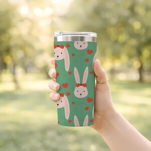 May include: A green travel tumbler featuring a repeating pattern of white bunny faces with pink ears and red bows, interspersed with red hearts. The tumbler is held by a hand, and the background is blurred green foliage.