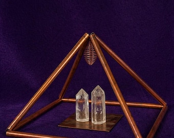 Puede incluir: Una estructura piramidal de cobre con dos puntas de cristal de cuarzo transparente sobre una base cuadrada de color cobre. La pirámide tiene una jaula de alambre colgando de la parte superior. El fondo es morado oscuro.