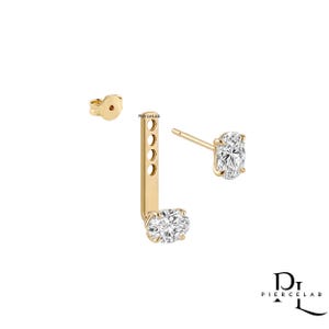 May include: A gold ear jacket earring featuring two oval-cut clear stones. One stone is set on a post, while the other is attached to a vertical bar with four adjustment holes. A separate gold earring back is included. The text PierceLab is visible.