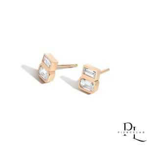 May include: A pair of gold stud earrings, each featuring two rectangular, clear gemstones. The earrings have a modern, geometric design and are set in a polished gold setting. The earrings are displayed on a white background.