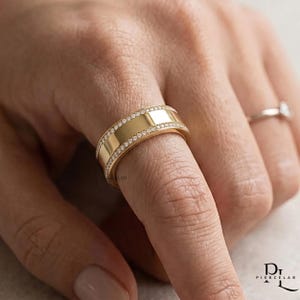 Könnte beinhalten: Ein breiter Goldring mit einer polierten, glatten Mitteloberfläche, die von zwei Reihen kleiner, klarer Pavé-gefasster Steine gesäumt ist. Der Ring wird an einem Finger getragen, wobei ein zweiter, dünnerer Goldring mit einem Stein sichtbar ist.