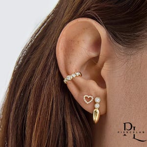 Puede incluir: Juego de aretes dorados. Incluye un brazalete de oreja curvado con cinco piedras transparentes, un arete en forma de corazón y un arete colgante con dos piedras transparentes y una base en forma de lágrima. Las joyas se usan en una oreja.