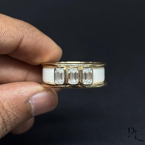 May include: A gold band ring featuring a central horizontal inlay of white material flanked by gold edges. Three clear, rectangular emerald-cut stones are set side-by-side in the center of the white inlay, creating a linear, multi-stone design.