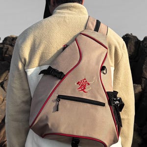 May include: A beige triangular sling backpack featuring red piping, a black zippered front pocket, and a black side mesh pocket. The front of the bag displays a red embroidered Japanese kanji character for love, with a thin red thread accent.