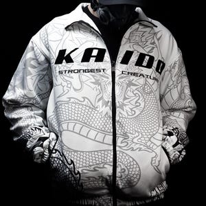 Könnte beinhalten: Eine weiße Reißverschlussjacke mit einer großen grauen Drachenillustration auf der Vorderseite und den Ärmeln. Der Text KAIDO ist in fetten schwarzen Buchstaben auf der Brust gedruckt, darunter die Worte STRONGEST CREATURE in kleinerer Schrift.