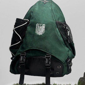 Può includere: Uno zaino verde scuro con un emblema ricamato in bianco delle Ali della Libertà sul davanti. La borsa presenta tasche laterali in rete nera, cinghie nere regolabili con fibbie in plastica e un ornamento a forma di croce color argento in alto.