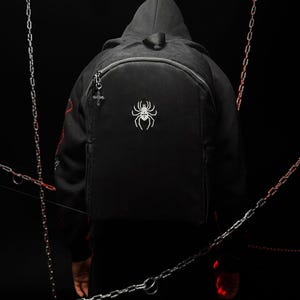 Puede incluir: Una mochila negra con un gráfico de araña bordado en blanco en el bolsillo delantero. El tirador de la cremallera está adornado con un pequeño dije de cruz plateado. La mochila tiene una parte superior redondeada, un compartimento principal y asa.