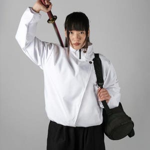 Könnte beinhalten: Eine weiße Jacke mit Stehkragen, diagonalem Frontverschluss und dunklen Knöpfen, kombiniert mit einer schwarzen Hose und einer schwarzen Umhängetasche. Das Outfit wird durch ein Katana mit rot umwickeltem Griff und goldfarbenem, rundem Stichblatt ergänzt.
