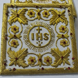 May include: A square white fabric corporal featuring intricate gold thread embroidery. The center displays the IHS monogram with a cross and three nails. The border and corners are decorated with gold floral motifs and small red and clear rhinestones.