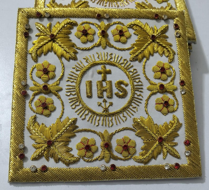 May include: A square white fabric corporal featuring intricate gold thread embroidery. The center displays the IHS monogram with a cross and three nails. The border and corners are decorated with gold floral motifs and small red and clear rhinestones.