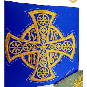 Könnte beinhalten: Ein goldgesticktes Kreuzdesign auf blauem Stoff. Das Kreuz weist komplizierte Blumen- und geometrische Muster mit kyrillischen Buchstaben auf. Die Stickerei ist detailliert und scheint handgenäht zu sein, was ein traditionelles Handwerk zeigt.