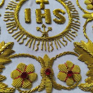 May include: A white fabric panel featuring intricate gold metallic thread embroidery. The design includes the letters IHS topped with a cross inside a circular border, surrounded by stylized gold floral motifs and small embedded red and clear crystals.