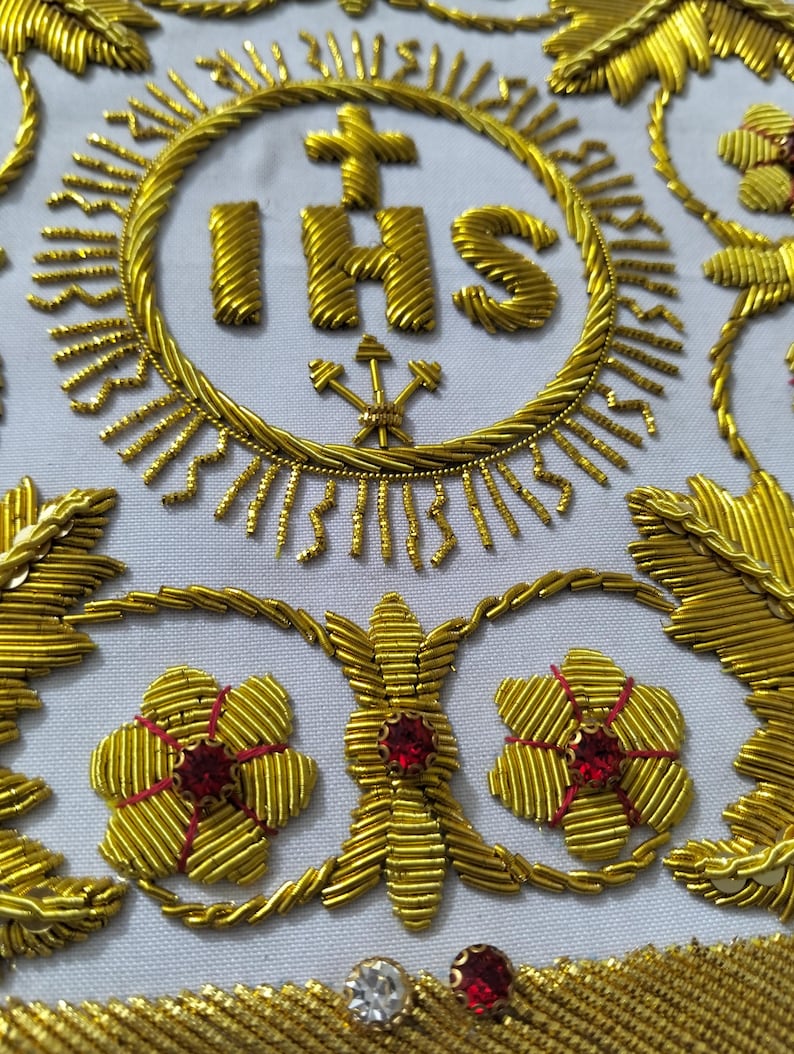 May include: A white fabric panel featuring intricate gold metallic thread embroidery. The design includes the letters IHS topped with a cross inside a circular border, surrounded by stylized gold floral motifs and small embedded red and clear crystals.