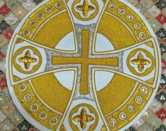 May include: Circular religious emblem with a gold cross design on a white background. The cross is surrounded by gold embroidery and smaller crosses at the corners. The design is on a patterned fabric.
