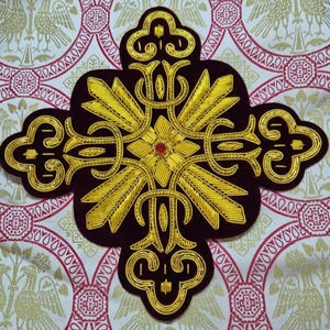 Puede incluir: Aplicación decorativa en forma de cruz, con bordado de hilo dorado sobre una base burdeos oscuro. Presenta una pequeña gema roja en el centro. El fondo es una tela estampada con detalles en rojo y dorado.