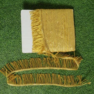 May include: A roll of gold fringe trim with a white cardboard backing. Two lengths of the fringe are unrolled, showing the detailed texture. The trim is on a green surface.