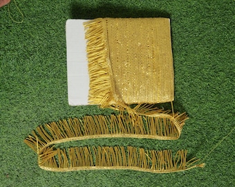 May include: A roll of gold fringe trim with a white cardboard backing. Two lengths of the fringe are unrolled, showing the detailed texture. The trim is on a green surface.