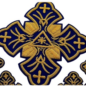 Może przedstawiać: Haftowana granatowa naszywka ze złotymi nićmi. Projekt przedstawia kształt krzyża ze złotymi detalami, centralny trójkąt z okiem i krzyże na rogach. Naszywka ma bogaty, dekoracyjny styl.