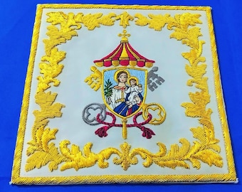 May include: A square white fabric panel featuring intricate gold thread embroidery along the border. The center displays a colorful embroidered crest with a figure holding a child, topped by a red and gold canopy, flanked by two keys.