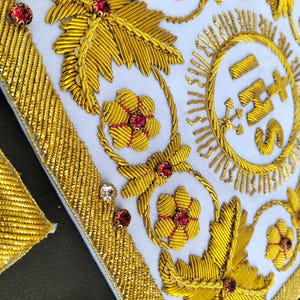 May include: A white fabric panel featuring intricate gold metallic thread embroidery in floral and leaf patterns. The design includes the letters IHS centered within a circular sunburst motif, accented with small red and clear faceted crystals.