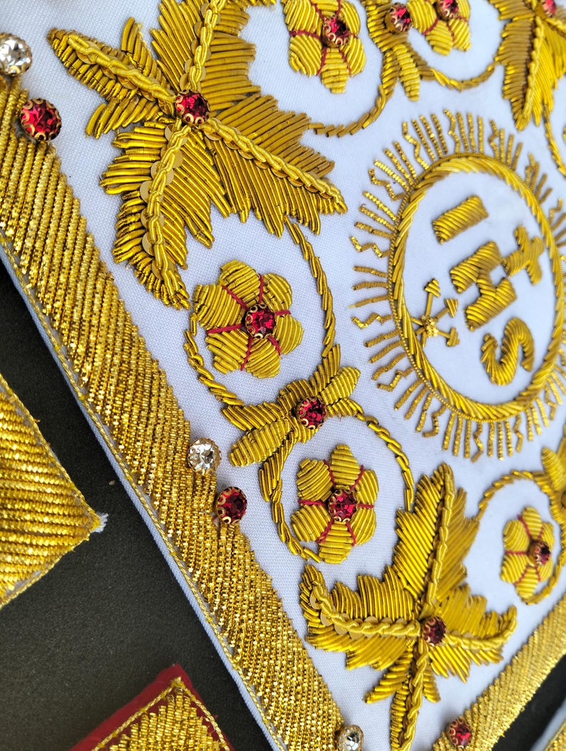 May include: A white fabric panel featuring intricate gold metallic thread embroidery in floral and leaf patterns. The design includes the letters IHS centered within a circular sunburst motif, accented with small red and clear faceted crystals.